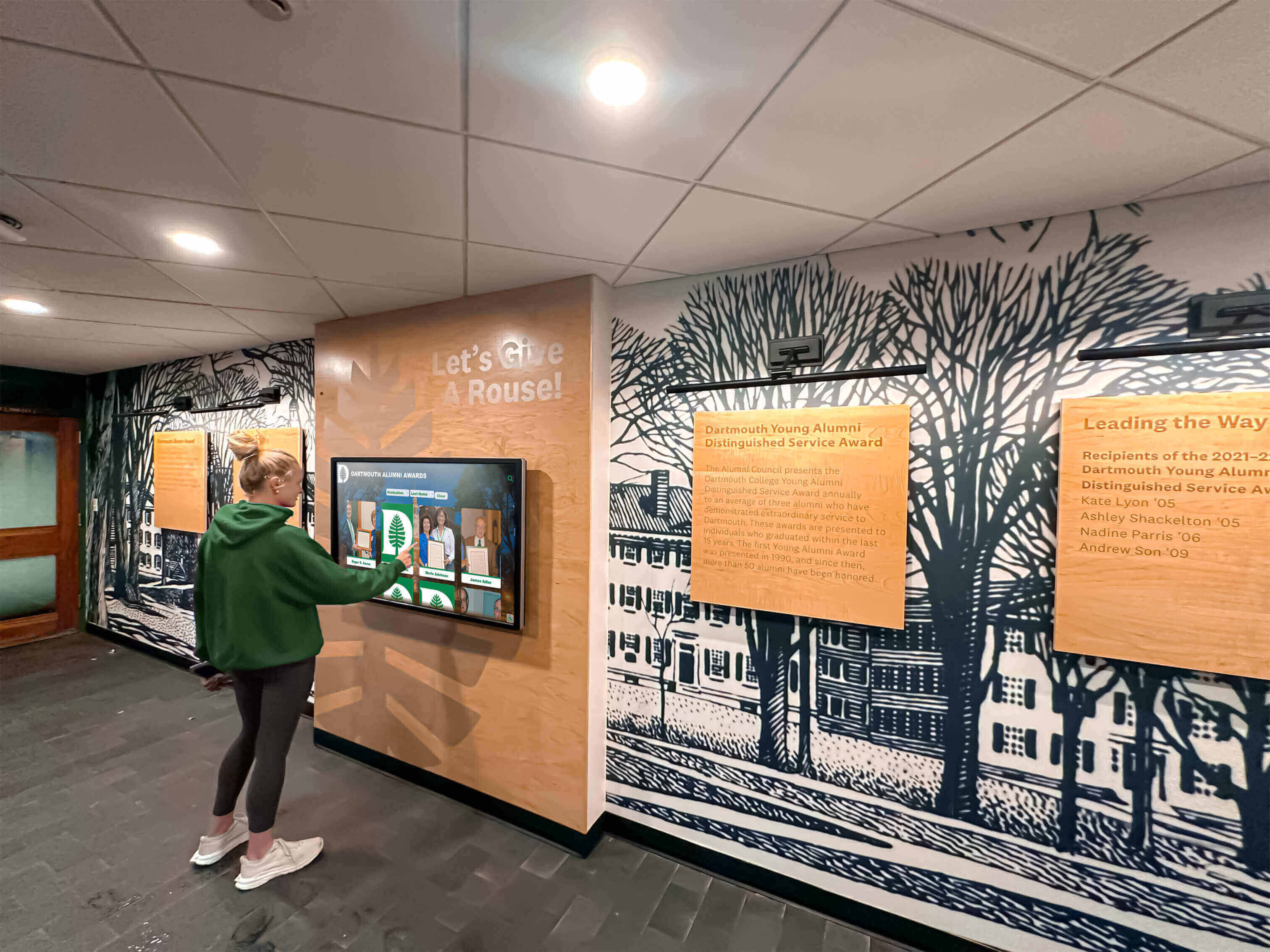 High school student in green hoodie browsing inductee profiles on a touchscreen display mounted in a school alumni recognition hallway Student in green hoodie using a touchscreen hall of fame display in an alumni hallway